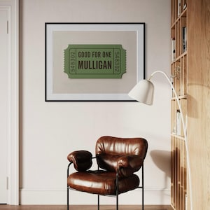 May include: A framed print featuring a green ticket design with the text "GOOD FOR ONE MULLIGAN." The print is set against a neutral background and framed in black. A brown leather armchair with black metal legs sits below the artwork.