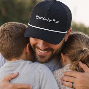 Può includere: Cappellino da baseball nero con la scritta "Best Dad by Par" in corsivo bianco. Il cappellino ha un dettaglio di corda bianca lungo il bordo. L'immagine mostra una persona che indossa il cappellino, che abbraccia due bambini.