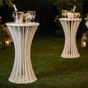 May include: Three white pedestal tables with curved, vertical supports stand on a lawn. Each table displays items such as champagne, glasses, flowers, and canapés. The scene is illuminated by soft outdoor lighting, creating an elegant atmosphere.
