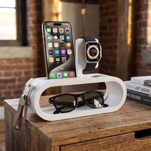 May include: A white charging station, oval in shape, holds a smartphone, smartwatch, and sunglasses. The phone displays app icons, and the watch has a black strap. Black sunglasses rest in the lower section. Keys hang on the side.