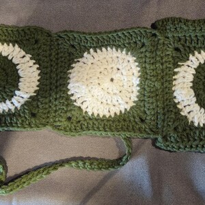 May include: A handmade crochet piece consisting of three connected square panels in dark olive green yarn. The two outer squares feature white crescent moon motifs, while the center square displays a white circular sun design. A long green cord extends from the bottom.