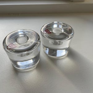 Peut inclure: Deux bougeoirs en verre transparent de forme arrondie. Chaque bougeoir a une ouverture circulaire en haut et une base épaisse. De petites étiquettes sont visibles sur le côté de chaque bougeoir. Les bougeoirs sont sur une surface blanche.