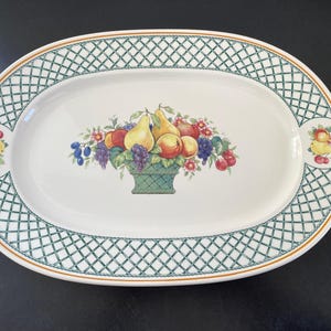 May include: Oval serving platter with a white surface and a green lattice border. The center features a basket of colorful fruit including pears, apples, grapes, and cherries. A decorative piece for serving.