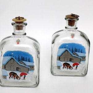 Könnte beinhalten: Zwei klare Glasflaschen mit einer Winterlandschaft bemalt. Die Szene zeigt eine verschneite Landschaft mit einem Haus, Bäumen und Figuren. Jede Flasche hat einen Korken mit einem Metallring. Die Flaschen haben eine quadratische Form.