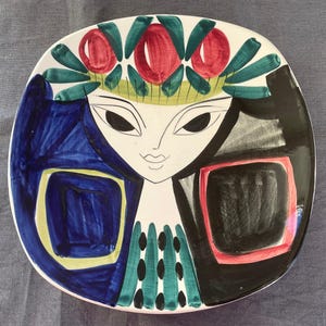以下が含まれることがあります： 正方形のセラミックプレート、様式化された顔のデザイン。 プレートは、黒で縁取られた顔、青、赤、緑のアクセントを備えた白い背景が特徴です。 デザインには、赤い花と緑の葉の王冠が含まれています。