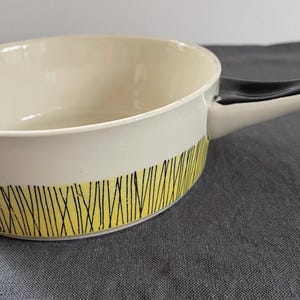 Può includere: Un pentolino in ceramica con interno ed esterno color crema. La parte inferiore dell'esterno presenta una fascia gialla decorata con un motivo di sottili linee nere verticali. Include un lungo manico nero ricurvo fissato sul lato.