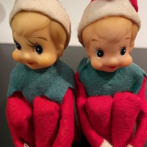 Peut inclure: Deux figurines d'elfes de Noël vintage. Chaque elfe a un visage en plastique, des cheveux blonds et porte une tenue en feutre rouge avec un col vert et un chapeau rouge et blanc. Les elfes sont assis, les jambes pliées.