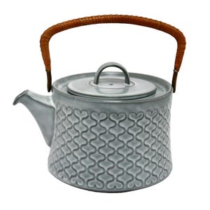 Può includere: Una teiera in ceramica grigia con un manico intrecciato marrone e un motivo a forma di cuore. La teiera ha un coperchio e un beccuccio.