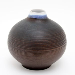 Peut inclure: Un vase rond en céramique fait à la main, avec un extérieur brun foncé texturé. Le vase a un intérieur blanc, une bande bleue au col et une base large et stable. Le vase est idéal pour exposer des fleurs ou comme élément décoratif.