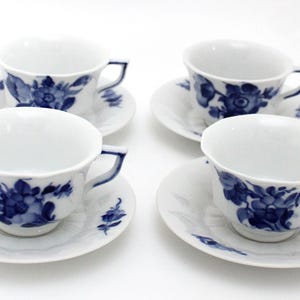 Op de afbeelding: Vier witte porseleinen kopjes en schotels. Elke kop en schotel heeft een blauw bloemmotief. De kopjes hebben een gebogen oor en de schotels zijn rond. Een klassieke theeset.