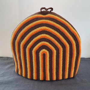 Könnte beinhalten: Eine gehäkelte Teewärmer im Retro-Stil. Der Teewärmer hat ein sich wiederholendes Muster aus braunen, orangefarbenen und gelben horizontalen Streifen. Oben befindet sich eine kleine Schlaufe zum einfachen Anfassen. Der Teewärmer ist für eine Teekanne konzipiert.