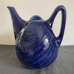 May include: A deep cobalt blue ceramic teapot featuring a raised, textured herringbone or knit-like pattern across its rounded body. The teapot includes a curved handle, a pointed spout, and a matching lid with a small, rounded knob on top.