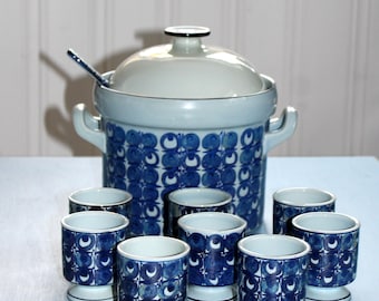 May include: A vintage ceramic serving set featuring a large lidded soup tureen with handles and a ladle, along with eight matching egg cups. The set is white and blue, with a repeating floral pattern. The egg cups have a pedestal base.