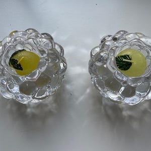 Può includere: Due portacandele in vetro trasparente con un design a bolle. Ogni portacandele contiene una candela gialla con un disegno a foglia verde. I portacandele sono su una superficie bianca.