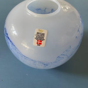 Op de afbeelding: Een ronde, lichtblauwe glazen vaas met een brede opening. De vaas heeft een gemarmerd ontwerp met donkerdere blauwe wervelingen. Een sticker op de voorkant leest "HOLMEGAARD COPENHAGEN". De vaas staat op een lichtblauwe ondergrond.