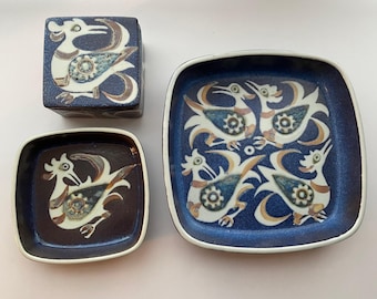 Könnte beinhalten: Ein Set aus drei Keramikteilen: eine kleine quadratische Box mit Deckel und zwei quadratische Schalen. Jedes Stück zeigt ein stilisiertes Vogeldesign in Weiß, Braun und Blau auf einem dunkelblauen Hintergrund. Das Set ist wahrscheinlich für dekorative oder servierende Zwecke.