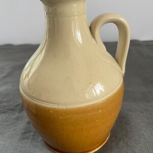 May include: A two-tone ceramic jug with a handle. The upper half is a light beige colour, and the lower half is a warm brown. The jug has a rounded body and a slightly flared spout. The handle is curved and attached to the side of the jug.