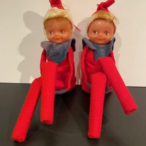 May include: Two vintage elf dolls with blonde hair, rosy cheeks, and blue eyes. They wear red felt outfits with matching hats and long red legs. Each doll has a grey collar.