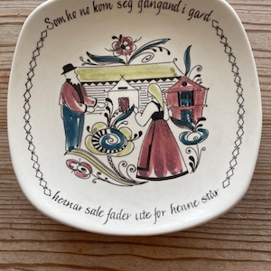 May include: A white ceramic plate with a square shape and a colourful folk art design. The plate features a couple standing in front of a house with a red roof. The text on the plate reads "Som ho no kom seg gangand i gard heinar sale fader lute for henne står."