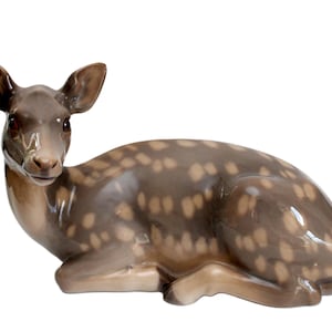 May include: A ceramic deer figurine in a reclining position. The deer features a brown coat with light brown spots. Its ears are erect, and its eyes are open. The figurine is a decorative item, suitable for display.