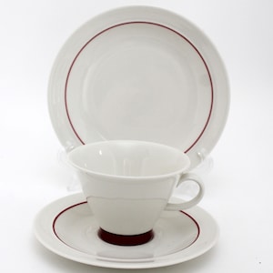 Puede incluir: Juego de té de porcelana blanca compuesto por taza, platillo y plato. Cada pieza tiene un borde fino de color rojo oscuro. La taza se apoya en el platillo, con un plato más grande detrás. El juego está diseñado para servir té o café.