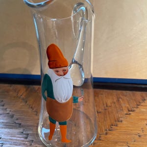 Peut inclure: Un pichet en verre transparent avec une anse, illustrant un gnome peint à la main portant un chapeau orange, une chemise verte, un tablier marron et des bottes orange. Le gnome tient une petite plante verte.
