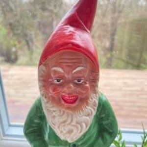 Könnte beinhalten: Eine Vintage-Garten-Zwergenfigur mit roter Mütze, grünem Hemd und weißem Bart. Der Zwerg hat ein lächelndes Gesicht mit einem schelmischen Ausdruck.