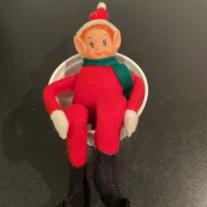 Può includere: Una bambola elfo vintage con corpo in feltro rosso, gambe in maglia nera e mani in feltro bianco. L'elfo indossa una sciarpa verde, un cappello rosso con finiture e pompon bianchi e ha un viso di plastica con occhi blu e un sorriso. La bambola è seduta in una ciotola bianca.