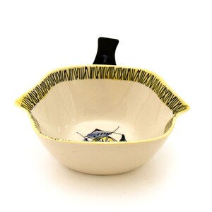 May include: Cream-colored ceramic bowl with a black handle and a yellow rim with black vertical lines. The bowl features a fish design in blue, yellow, and green. A decorative piece for serving snacks or displaying small items.