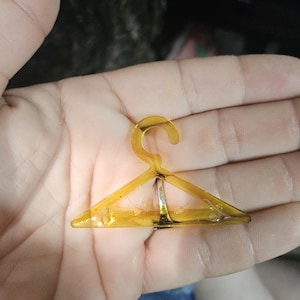 Peut inclure: Un petit cintre de vêtement miniature de couleur ambre transparente. Le cintre a une forme triangulaire avec un crochet incurvé en haut et un anneau métallique argenté au centre. Le cintre est tenu dans la main d'une personne.
