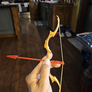 Puede incluir: Un arco y una flecha de juguete de plástico dorado. El arco está decorado con un patrón de espiral. La flecha es roja.