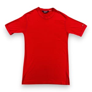 May include: A bright red t-shirt with short sleeves. The shirt has a crew neck and a small logo on the upper chest. A small red tag is visible on the left sleeve. The shirt is made of a soft material.