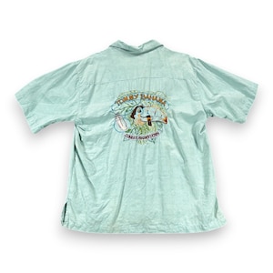 May include: Light blue short-sleeve button-down shirt. The back of the shirt features an embroidered design with a toucan, the words "Tommy Bahama," and "Jungle Shakedown." The shirt has a collared neckline and a relaxed fit.