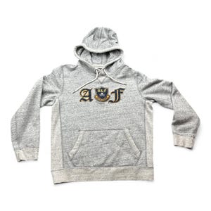 May include: A heather gray long-sleeve hooded sweatshirt featuring a front kangaroo pocket and a central embroidered crest logo with the letters A and F flanking a shield containing the text A&F and a small skull and crossbones graphic.
