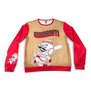 May include: A long-sleeved sweatshirt featuring the Cincinnati Reds baseball team logo. The shirt has a tan front with the word "Cincinnati" in red, and a baseball character. The sleeves are red.