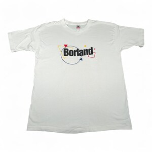 Op de afbeelding: Wit t-shirt met het woord "Borland" in zwart, met een geel, rood en blauw geometrisch ontwerp. Het shirt is gemaakt van een zacht materiaal en heeft korte mouwen.