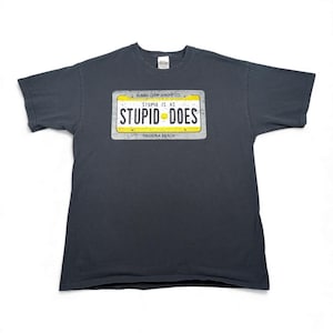 Könnte beinhalten: Dunkelgraues T-Shirt mit einem Nummernschild-Design mit der Aufschrift "STUPID IS AS STUPID DOES". Das Nummernschild hat einen gelben Hintergrund mit schwarzem Text und einem Smiley. Das Shirt trägt auch den Text "BUBBA GUMP SHRIMP CO." und "MADEIRA BEACH."