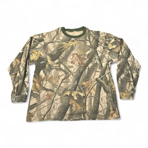 Könnte beinhalten: Langärmeliges Camouflage-Shirt mit Rundhalsausschnitt und grünem Kragen. Das Shirt hat ein Muster aus Braun, Beige und Grün, das einer Waldumgebung ähnelt. Die Ärmel sind lang und das Shirt ist aus weichem Material.