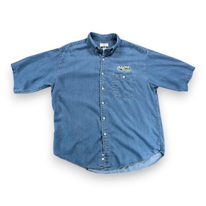 May include: A short-sleeve blue denim button-down shirt featuring a button-down collar and a single chest pocket. The shirt displays the text "SKOAL RACING" embroidered in white and green on the left chest area above the pocket.