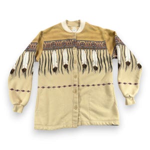 Könnte beinhalten: Beige Knopfjacke mit einem Design im indianischen Stil. Die Jacke hat einen beigen Körper, eine braune Passe und Ärmel mit einem Federmuster. Das Design umfasst weiße Federn mit braunen Spitzen und dekorative Verzierungen.