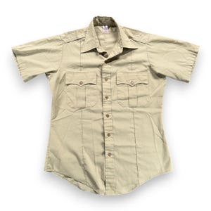 May include: A khaki-coloured short-sleeved shirt with two chest pockets and button fastenings. The shirt has a collared neckline and epaulettes on the shoulders. The shirt is made of a lightweight fabric.
