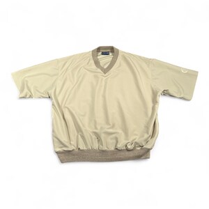 Puede incluir: Un cortavientos beige de manga corta con cuello en V y dobladillo acanalado. El cuello y el dobladillo son de un color beige más oscuro. Un pequeño logotipo es visible en la manga izquierda.