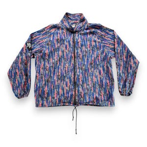 Può includere: Una giacca a maniche lunghe con zip e un motivo a pennellate verticali multicolore nei toni del blu, rosa, viola e grigio. La giacca presenta un colletto alto, una cerniera frontale nera, polsini elastici e un orlo con coulisse regolabile.