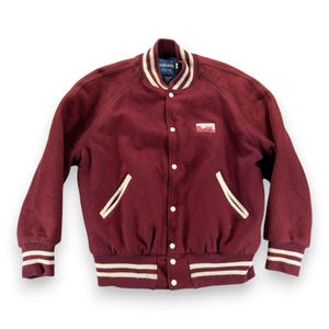 Puede incluir: Una chaqueta universitaria de color granate con cierre frontal de botones a presión, mangas largas y puños acanalados con rayas blancas. La chaqueta incluye dos bolsillos laterales con ribete blanco, cuello a rayas blancas y un parche.