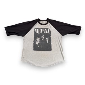 Peut inclure: T-shirt de style raglan gris avec des manches noires. Le t-shirt présente un graphisme noir et blanc du groupe Nirvana et le nom du groupe en lettres blanches en gras. Le t-shirt est de style vintage.
