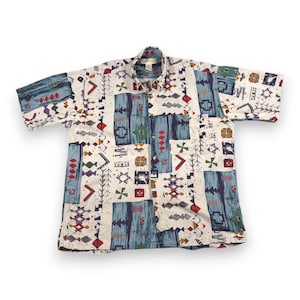 Puede incluir: Una camisa de manga corta con un colorido estampado geométrico. La camisa presenta un fondo blanco con formas azules, rojas, verdes y moradas. El cuello y los botones son visibles.