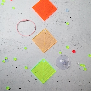 Könnte beinhalten: Drei rautenförmige Acrylornamente in Neonorange, Gelb und Limettengrün, jeweils mit einem Loch und eingravierten Blattadermustern. Ein rosa runder Kabelring und ein transparenter Saugnapfhaken sind im Lieferumfang der Ornamente enthalten.