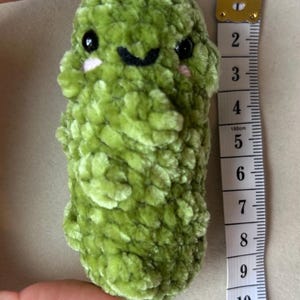 May include: A green plush pickle keyring featuring black safety eyes and embroidered pink cheeks. The textured yarn character is attached to a silver metal ring. A white measuring tape placed alongside shows the item is approximately 9 cm in length.