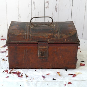 May include: A vintage, rectangular metal toolbox with a hinged lid and a handle. The toolbox is a rusty brown color with visible signs of wear and tear, including areas of rust and discoloration. The latch is a simple, rectangular design.