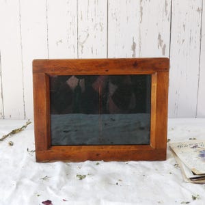 May include: A rectangular wooden frame with a dark interior, possibly for a picture or mirror. The frame is made of a warm-toned wood, and the glass or dark material inside is divided into two sections. The frame sits on a white surface.
